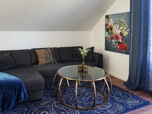 A living room with a dark grey sofa, a glass coffee table with a gold base, a blue patterned rug, and a floral painting on the wall. - 4 Person Holiday Home in Rimbo (Rånäs)