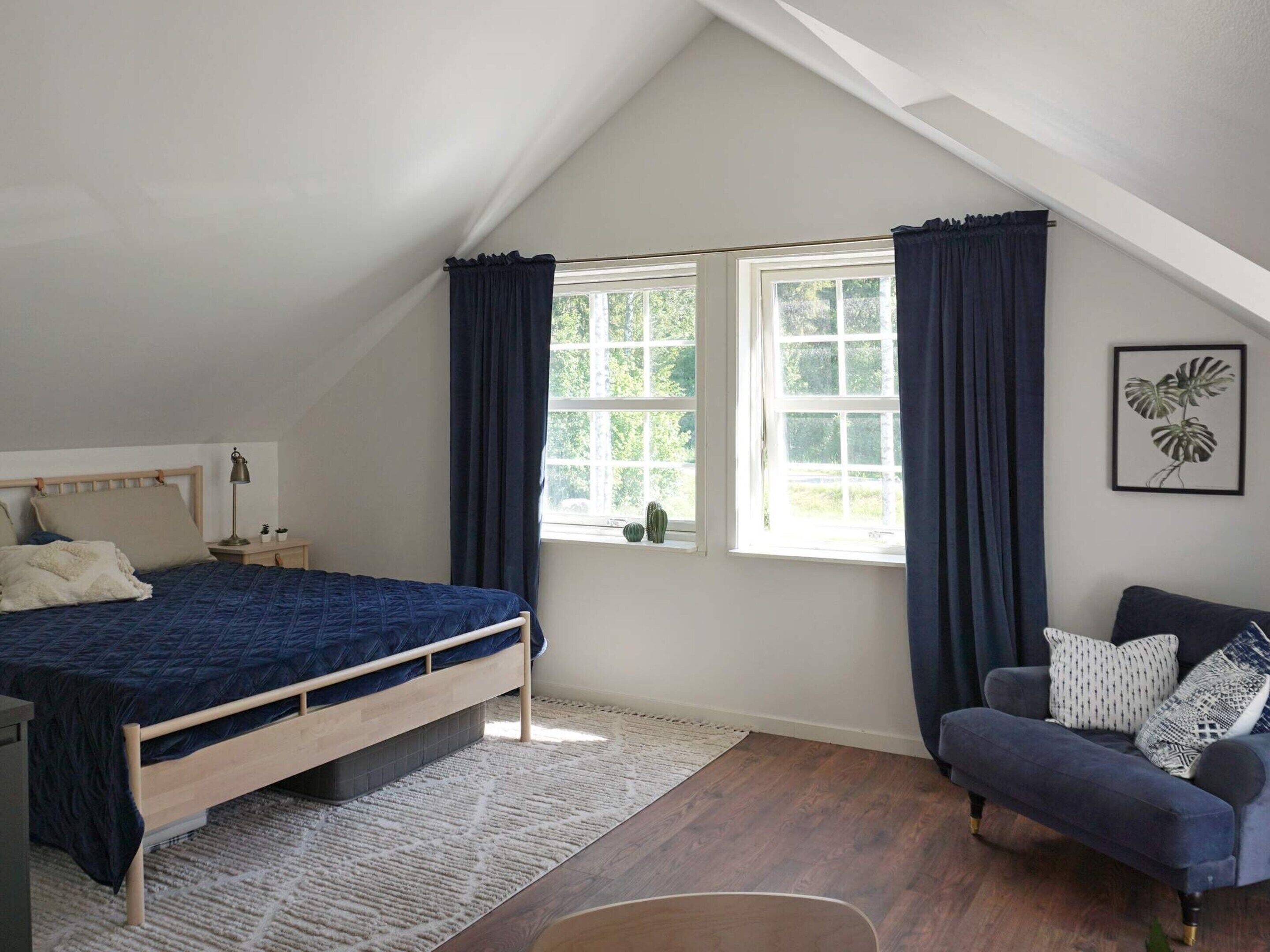 A bedroom with a bed, a chair, a window with curtains, a plant, and a framed picture on the wall.