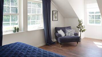 A bedroom with a large bed, a window with curtains, a dark-colored sofa, and a potted plant.