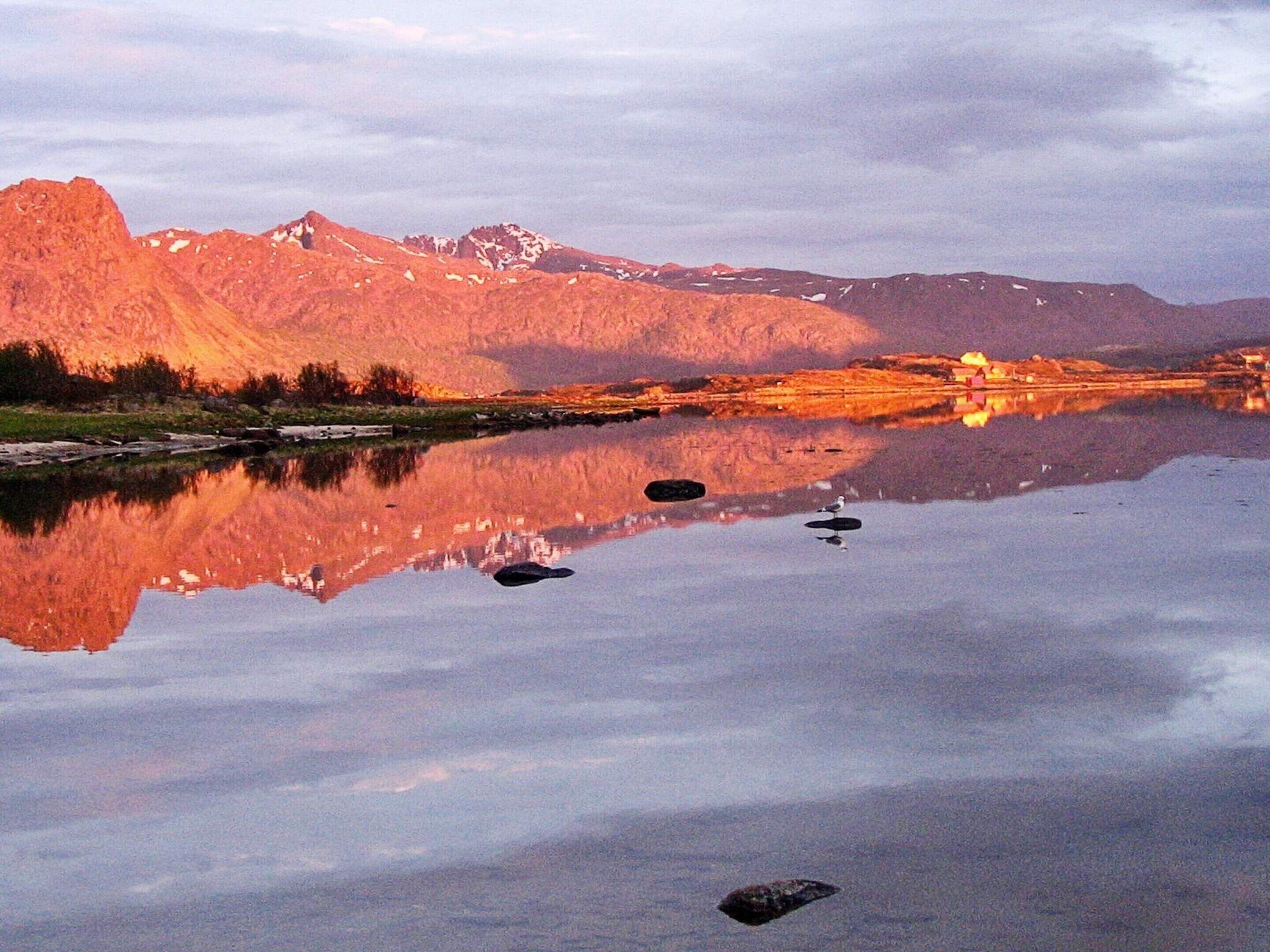 A calm lake reflecting mountains and a small building with lights, surrounded by a clear sky.
