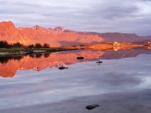 A calm lake reflecting mountains and a small building with lights, surrounded by a clear sky.