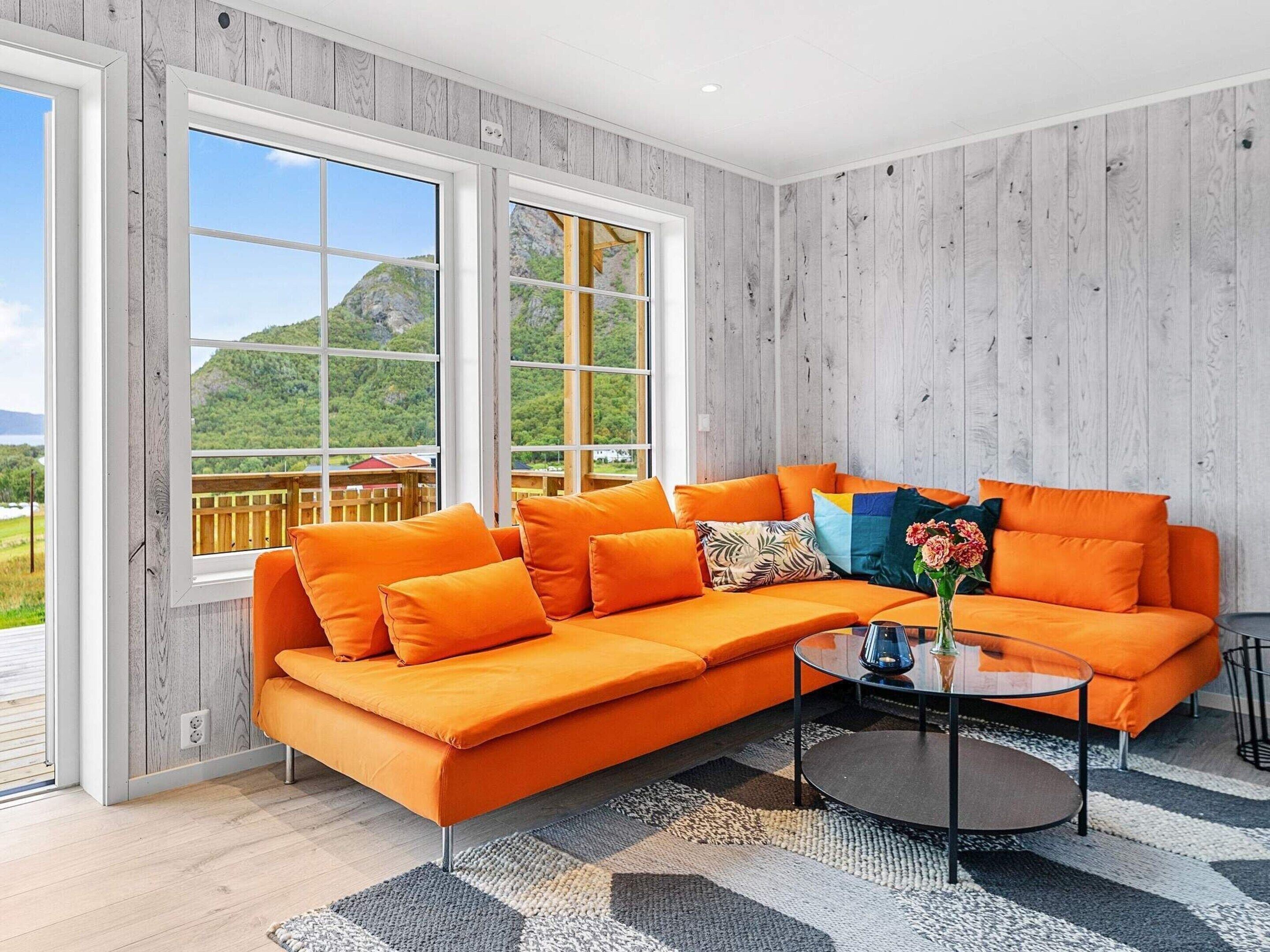 A modern living room with a large orange sectional sofa, a glass coffee table, and a view of a mountainous landscape through large windows.