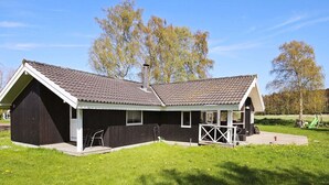 En liten, envåningsbyggnad med en mörk fasad och ett tak täckt med takpannor. Den har en täckt veranda med ett räcke och en liten utomhusplats med sittplatser.