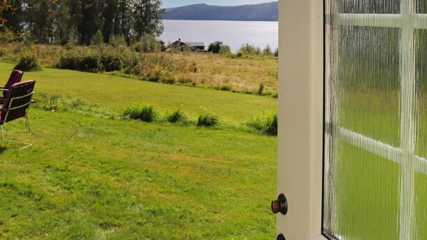 A view from a room with a large window overlooking a grassy field, a chair, and a distant body of water. - 6 Person Holiday Home in Ostra Antervik-by Traum (Östra Ämtervik)