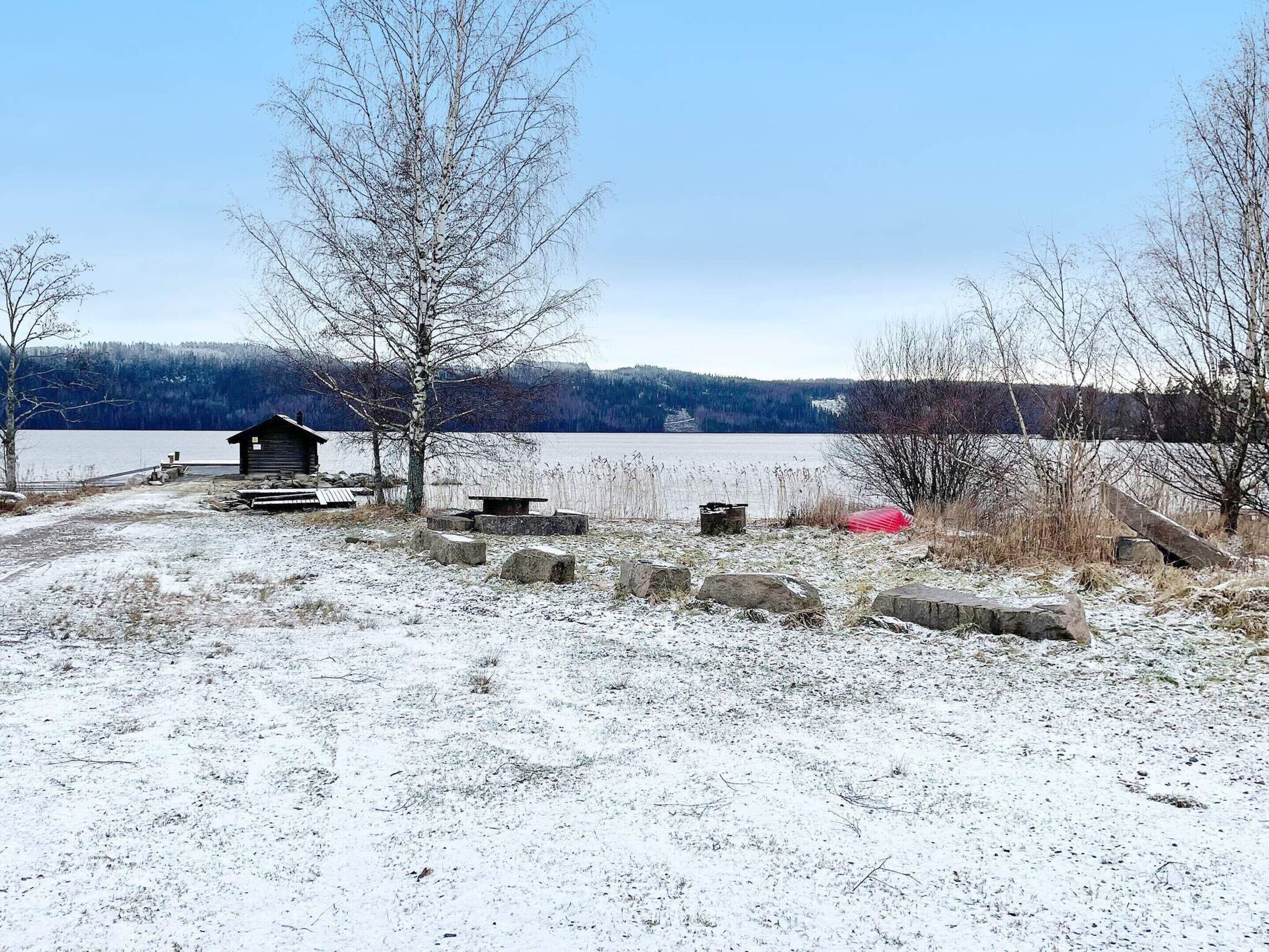 A cabin by a lake with a snow-covered ground, a small dock, and a bench.