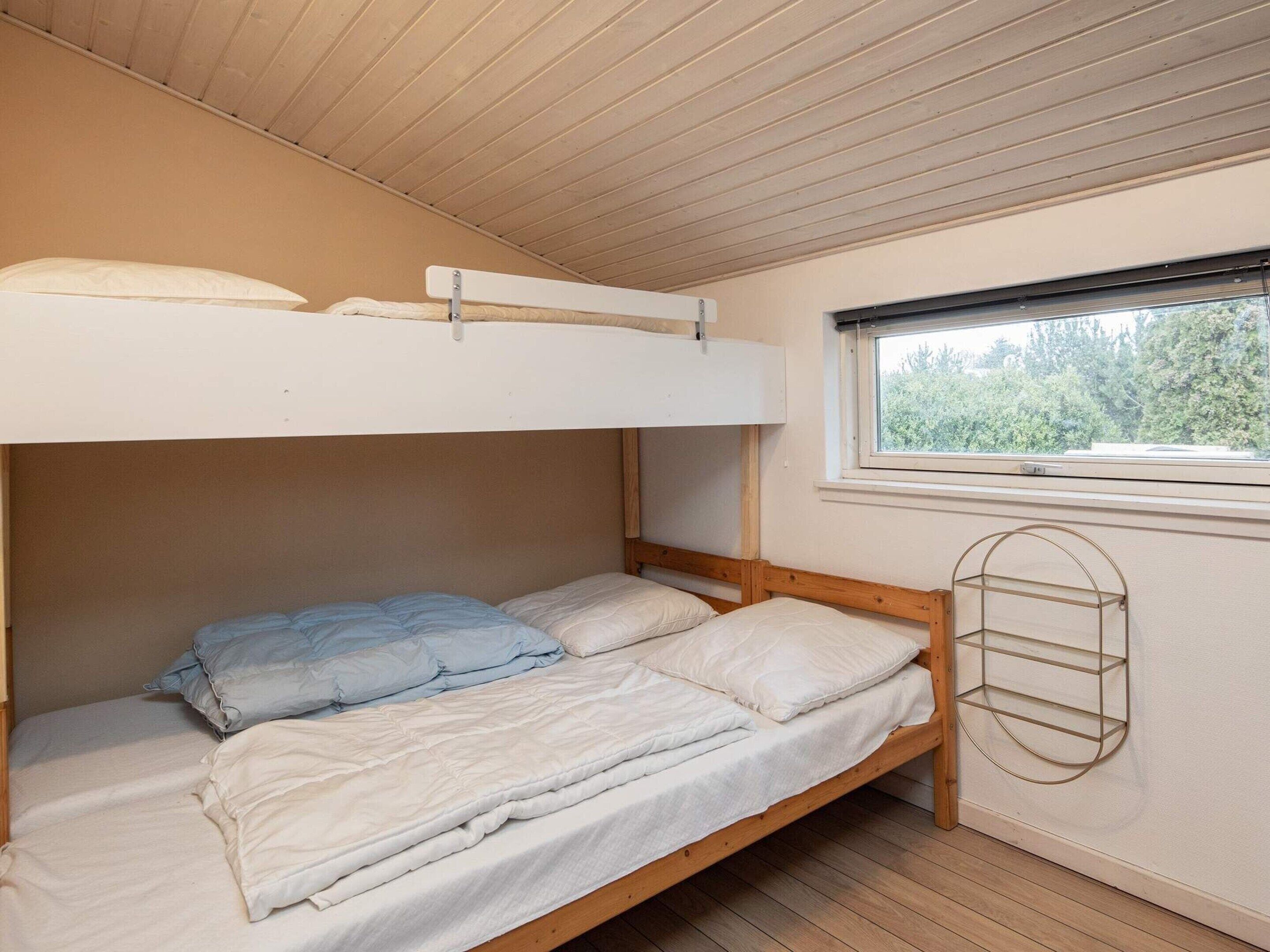 A bunk bed with a window above it, a wooden floor, and a metal shelf on the wall.