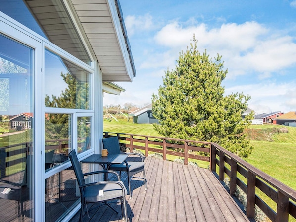 A wooden deck with outdoor furniture, a large window, and a view of a green landscape with trees and houses.