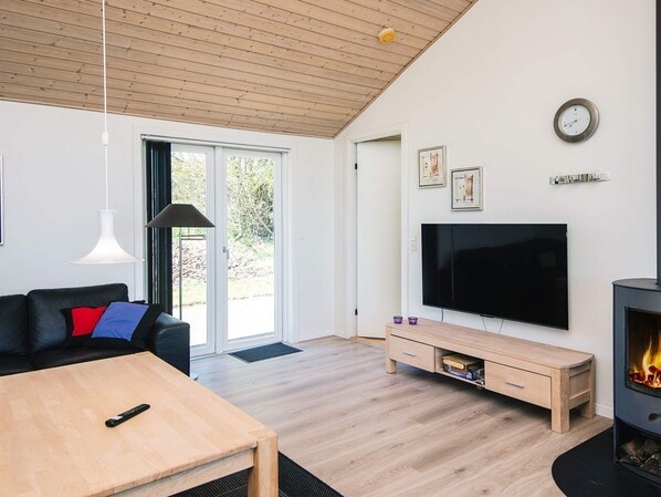 Una sala de estar moderna con techo de madera, un sofá negro con cojines rojos y azules, una mesa de centro de madera, un televisor sobre un mueble, una chimenea y una puerta que da a un balcón.