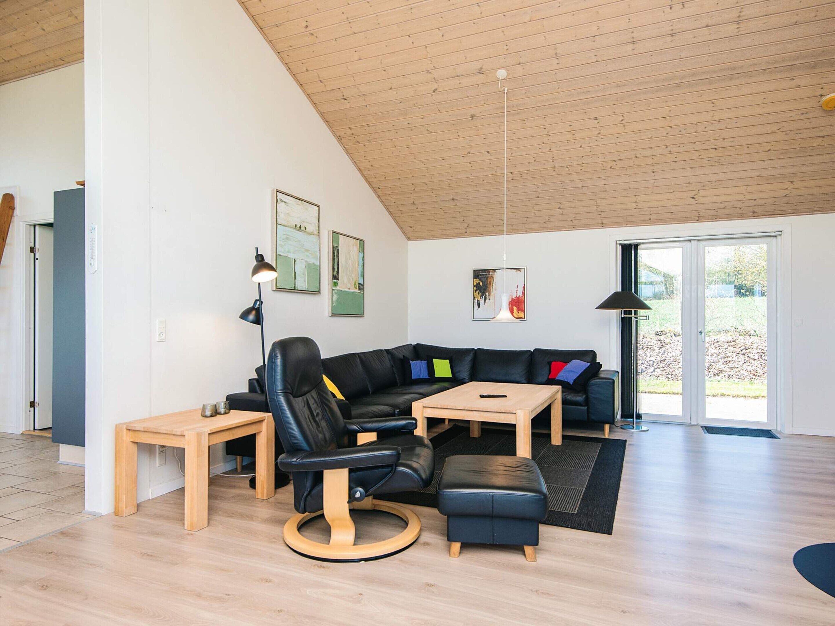A modern living room with a wooden ceiling, a black leather sofa, a wooden coffee table, and a black ottoman.
