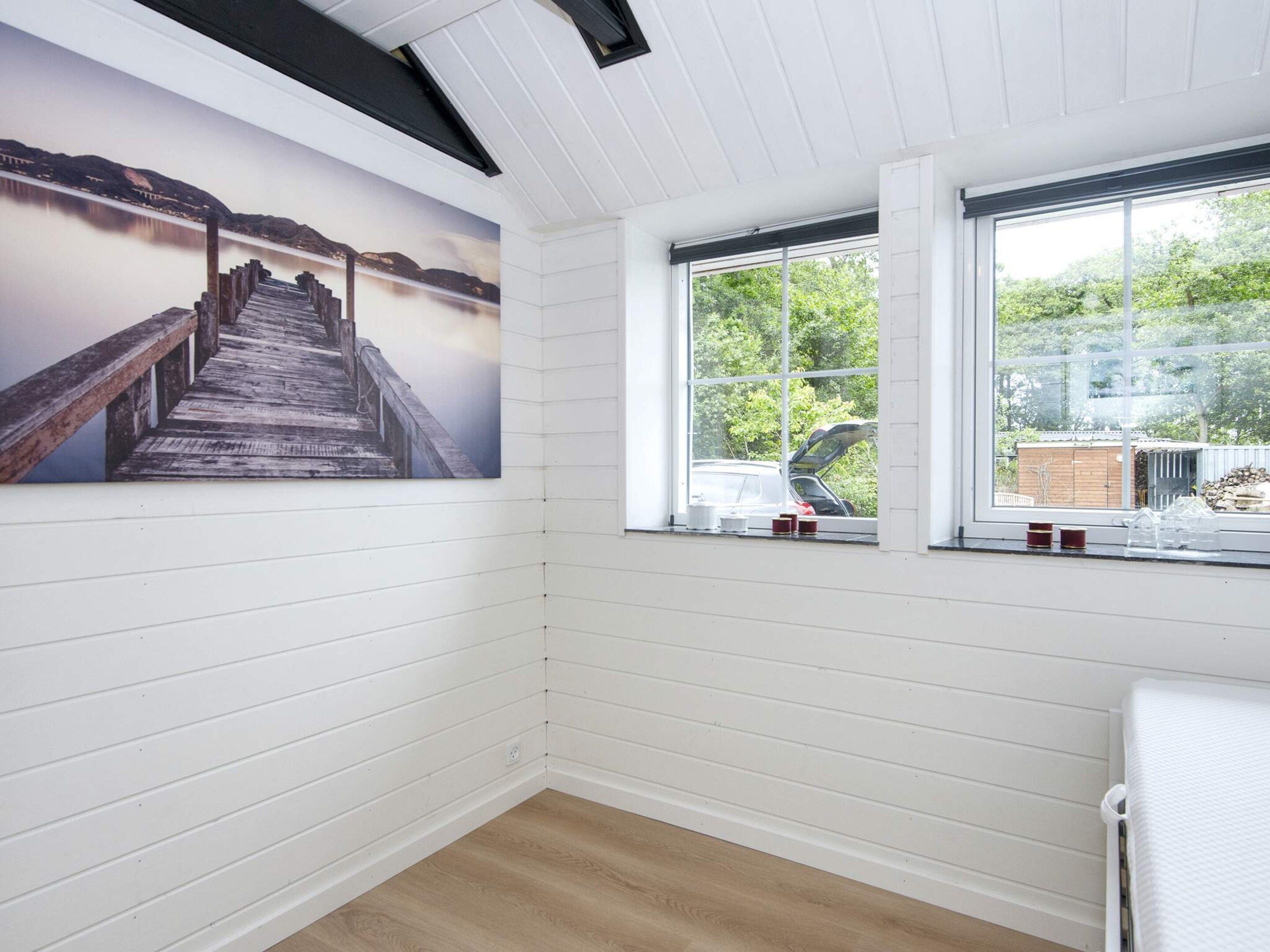 A room with white paneled walls, a wooden floor, a large window with a view of greenery, and a painting of a wooden pier.
