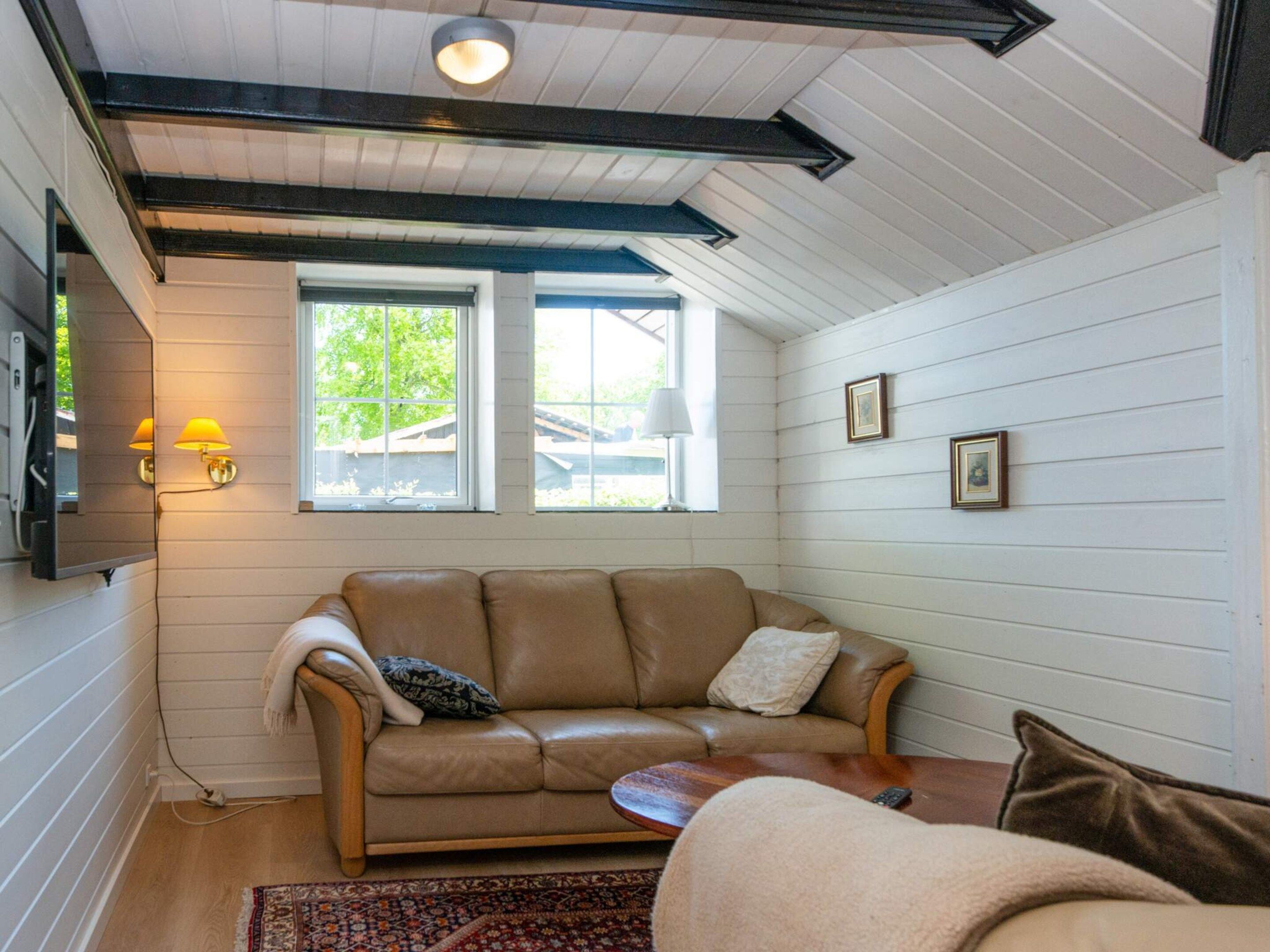 A cozy living room with a brown leather sofa, a small round coffee table, and a patterned rug. There are two windows providing natural light, and a flat-screen TV mounted on the wall.