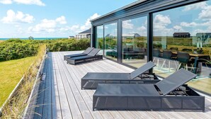 A modern outdoor deck with lounge chairs and a glass-enclosed dining area overlooking a coastal landscape. - 5 Star Holiday Home in Lokken (Lokken)