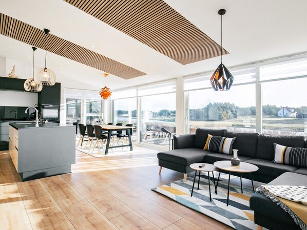 A modern kitchen with a central island, dining area with chairs, a sofa with cushions, and large windows offering a view.