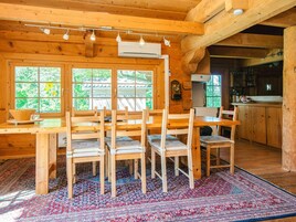 A wooden cabin interior with a dining area, kitchen, and large windows.