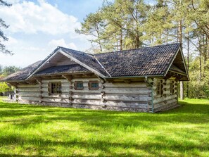 A log cabin with a steeply pitched roof and a stone foundation, surrounded by a grassy field and trees.
