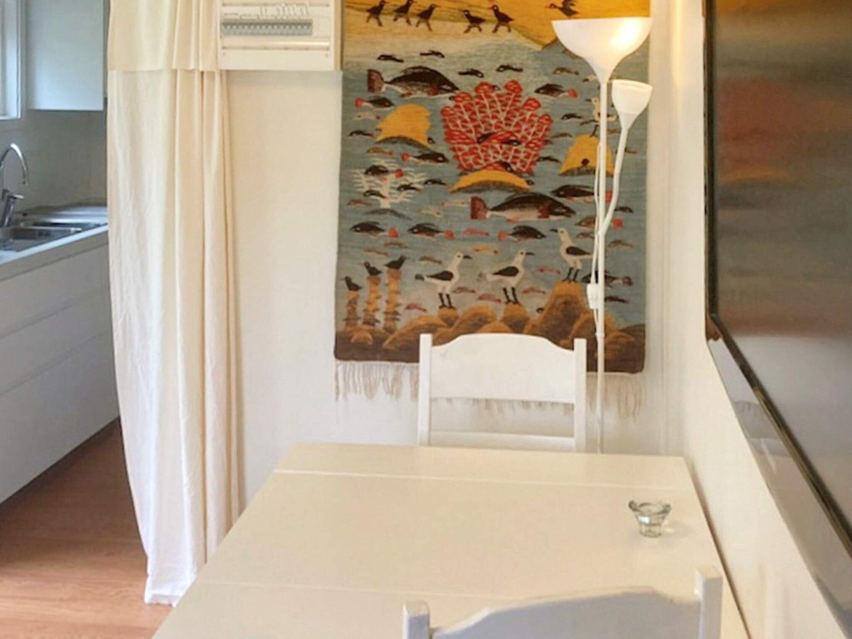 Une pièce avec une table blanche, une chaise blanche, une lampe, une tapisserie à motifs et un coin cuisine avec un évier.