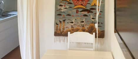 A room with a white table, a white chair, a lamp, a patterned tapestry, and a kitchen area with a sink.
