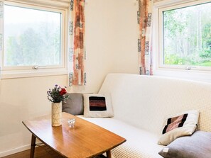 A room with a white sofa, a wooden table, and a vase with flowers. There are two windows with curtains.