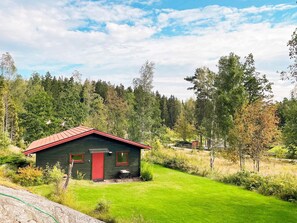 A small, dark green cabin with a red roof and two windows, surrounded by a well-maintained grassy area and dense forest.