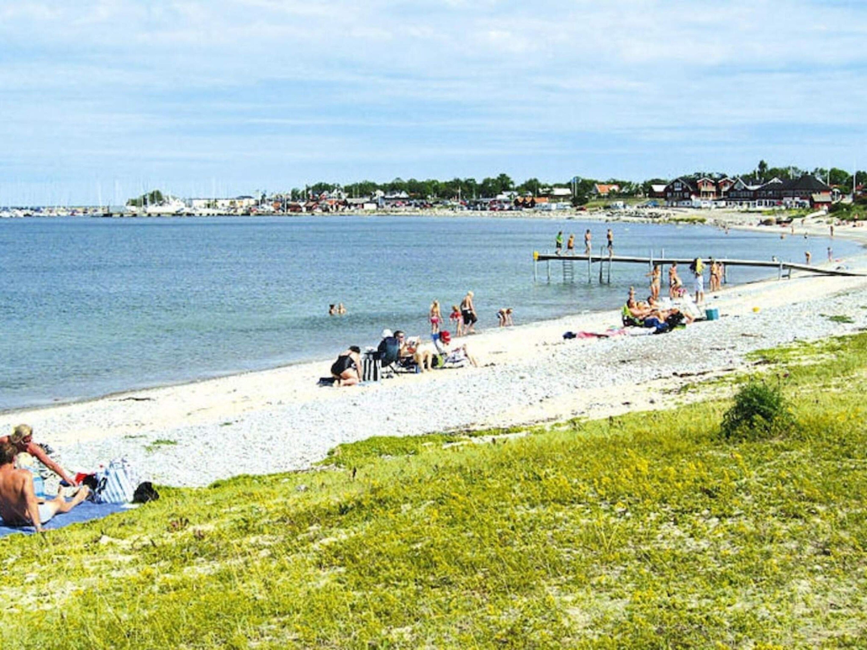 Ein Strand mit badenden und sonnenden Personen, ein Steg, der ins Wasser ragt, und Gebäude im Hintergrund.