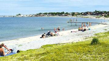 Ein Strand mit badenden und sonnenden Personen, ein Steg, der ins Wasser ragt, und Gebäude im Hintergrund.