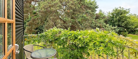 A wooden deck with two wicker chairs and a glass table, surrounded by a metal railing and lush greenery.