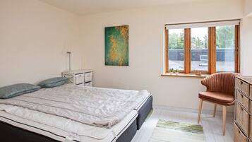 A bedroom with a bed, bedside table, lamp, artwork, window, chair, and storage unit.