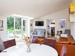 A modern living room with a sofa, a dining table with chairs, a flat-screen TV mounted on the wall, and a large window providing a view of the outdoors. - 4 Star Holiday Home in Vaeggerlose (Vaeggerlose)