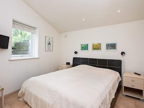 A bedroom with a bed, a nightstand, a bedside table, a TV mounted on the wall, and a window with a view of greenery. - 4 Star Holiday Home in Vaeggerlose (Vaeggerlose)