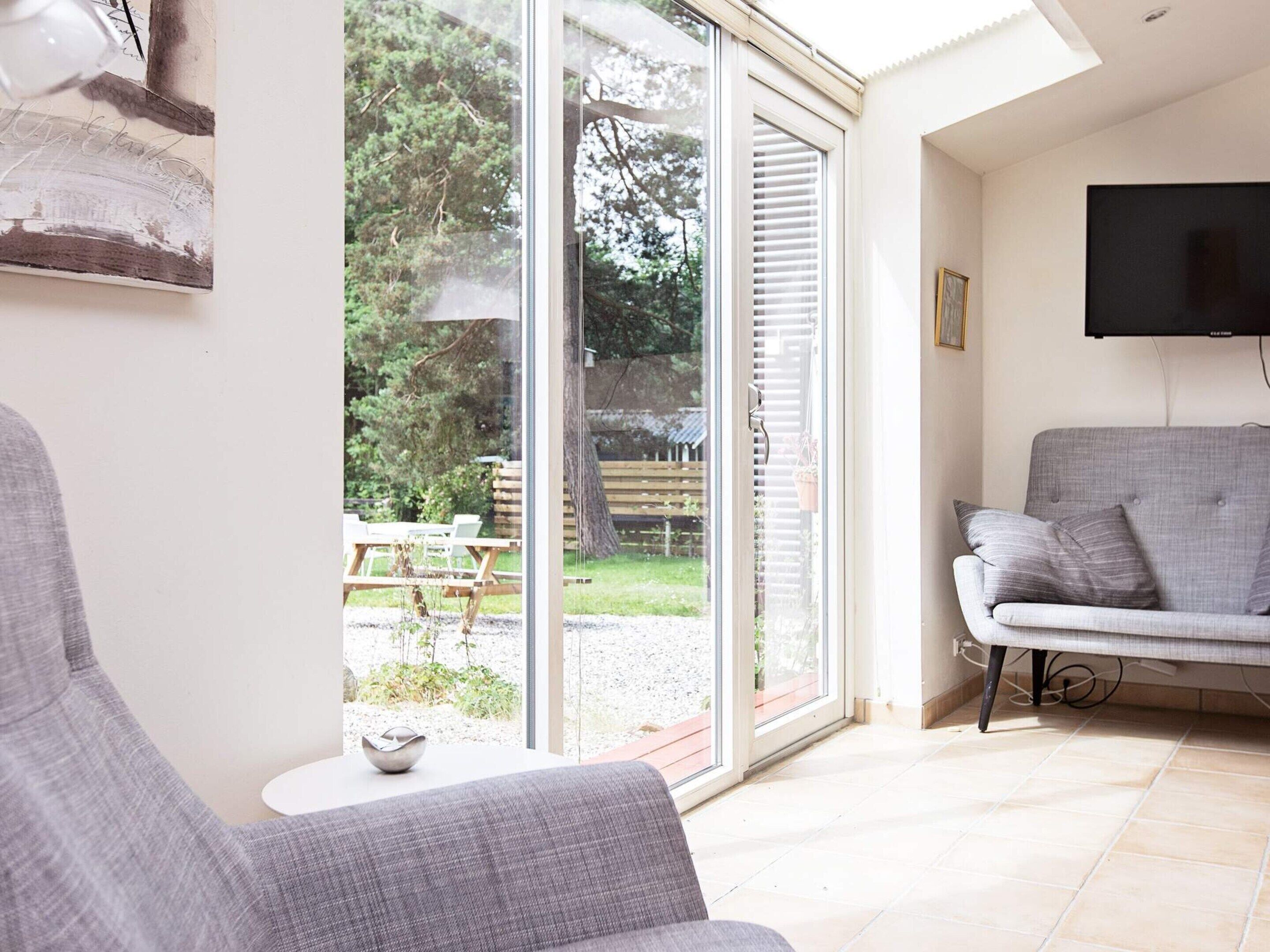 Ein Wohnzimmer mit grauem Sofa, einem flachbildfernseher an der Wand und einer Glastür, die zu einem Außenbereich mit Holzterrasse und Grünflächen führt.