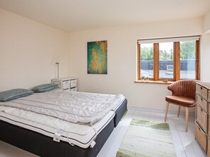 A bedroom with a bed, bedside table, lamp, artwork, window, chair, and storage unit.