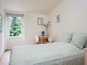 A bedroom with a bed, a desk, two chairs, a lamp, and a window with a view of trees.
