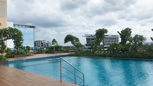 A swimming pool with a wooden deck and metal railings, surrounded by lush greenery and modern buildings in the background.