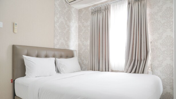 A neatly made bed with white linens in a room with patterned wallpaper and a large window with curtains. - Cozy And Well Designed 2Br At Green Pramuka City Apartment (Jakarta)