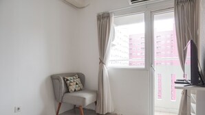 A room with a grey armchair, a patterned rug, a desk with a computer, and a view of a red building through a window with curtains. - Cozy And Well Designed 2Br At Green Pramuka City Apartment (Jakarta)