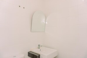 Une salle de bain avec des carreaux blancs, un lavabo rectangulaire surmonté d’un miroir et des toilettes situées à gauche.