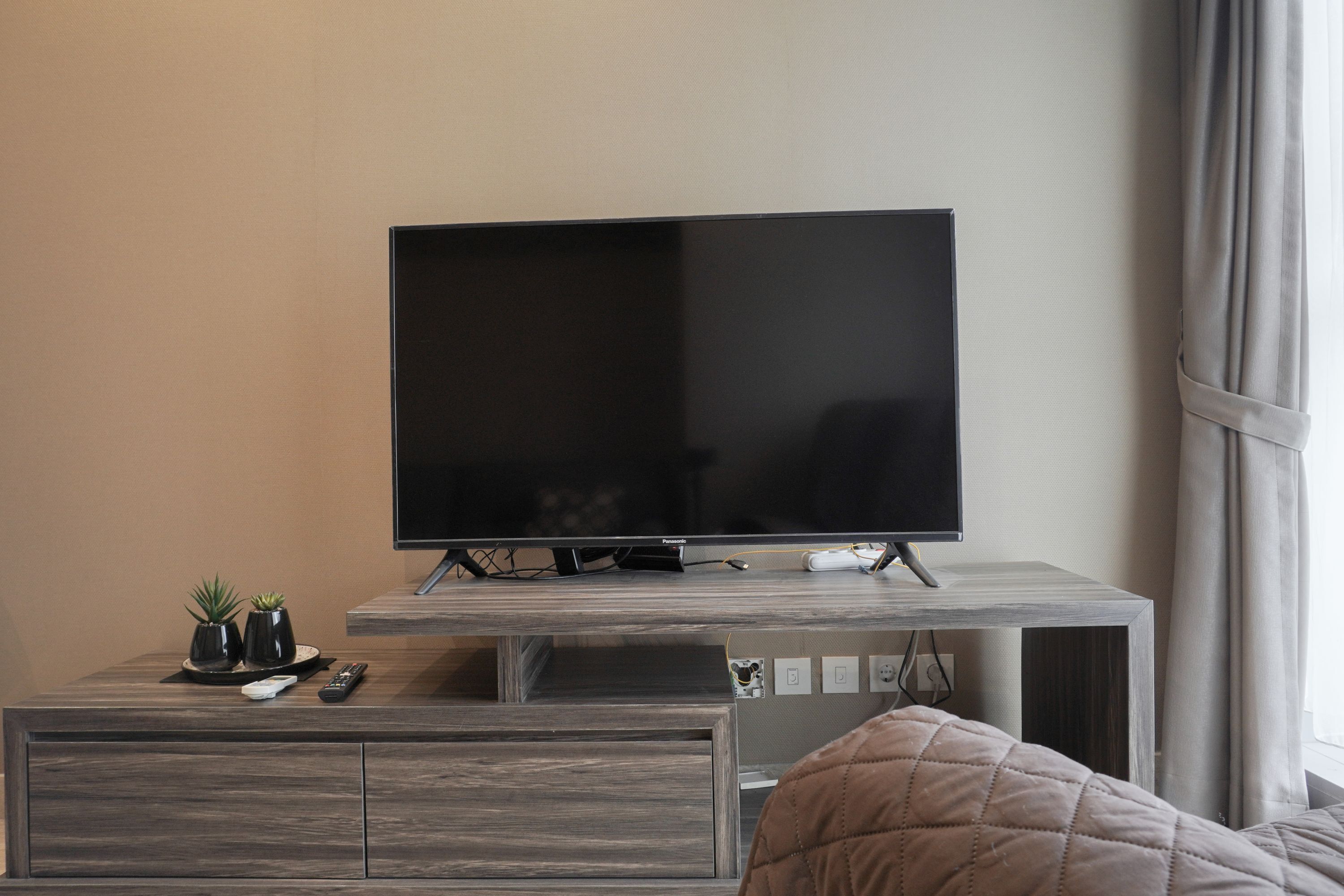 A modern living room with a flat-screen TV on a wooden stand, a small table with two potted plants, a remote control, and a bed with a beige quilt.