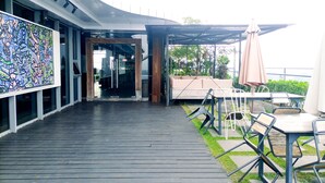 An outdoor patio area with a wooden deck, outdoor seating with tables and chairs, and a glass entrance to a building. - Cozy Studio Room At Uttara The Icon Apartment (Depok)