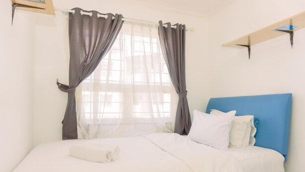 A neatly made bed with white linens and a blue headboard, a window with sheer curtains, and a white wall with a shelf. - Comfort 2Br At 22Nd Floor Menara Kebon Jeruk Apartment (Jakarta)