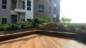 A rooftop terrace with a wooden floor, surrounded by greenery and a building with multiple windows and balconies. - Nice And Comfortable 2Br At 20Th Floor Tokyo Riverside Apartment (Teluknaga)
