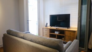 Un salón con un sofá gris, un televisor sobre un mueble de madera y un ventanal corredero que da a un balcón.