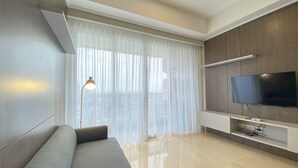 A modern living room with a sofa, a TV mounted on the wall, and a large window with sheer curtains. - Comfort 2Br At Menteng Park Apartment (Jakarta)