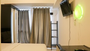 A hotel room with a bed, curtains, a TV mounted on the wall, and a desk with a remote control. - Comfy And Simply Studio Tokyo Riverside Pik 2 Apartment (Teluknaga)