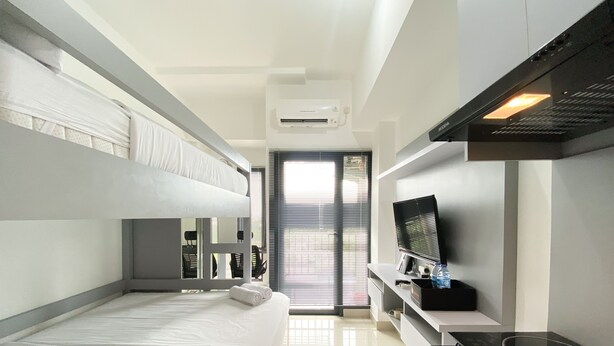 A modern hotel room with bunk beds, a flat-screen TV, a built-in stove, and a large window with blinds. - Cozy Stay Studio At Sayana Bekasi Apartment (Tarumajaya)