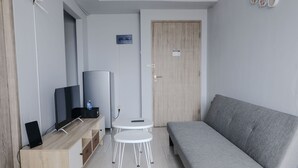 A modern, compact room with a grey sofa, a wooden TV stand, a flat-screen TV, a small white table, and a door. - Homey 1Br Apartment At Newton Residence Bandung (Bandung)