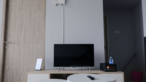 A modern hotel room with a wooden door, a flat-screen TV on a wooden stand, a small table with a remote control, and a water bottle on a side table. - Homey 1Br Apartment At Newton Residence Bandung (Bandung)