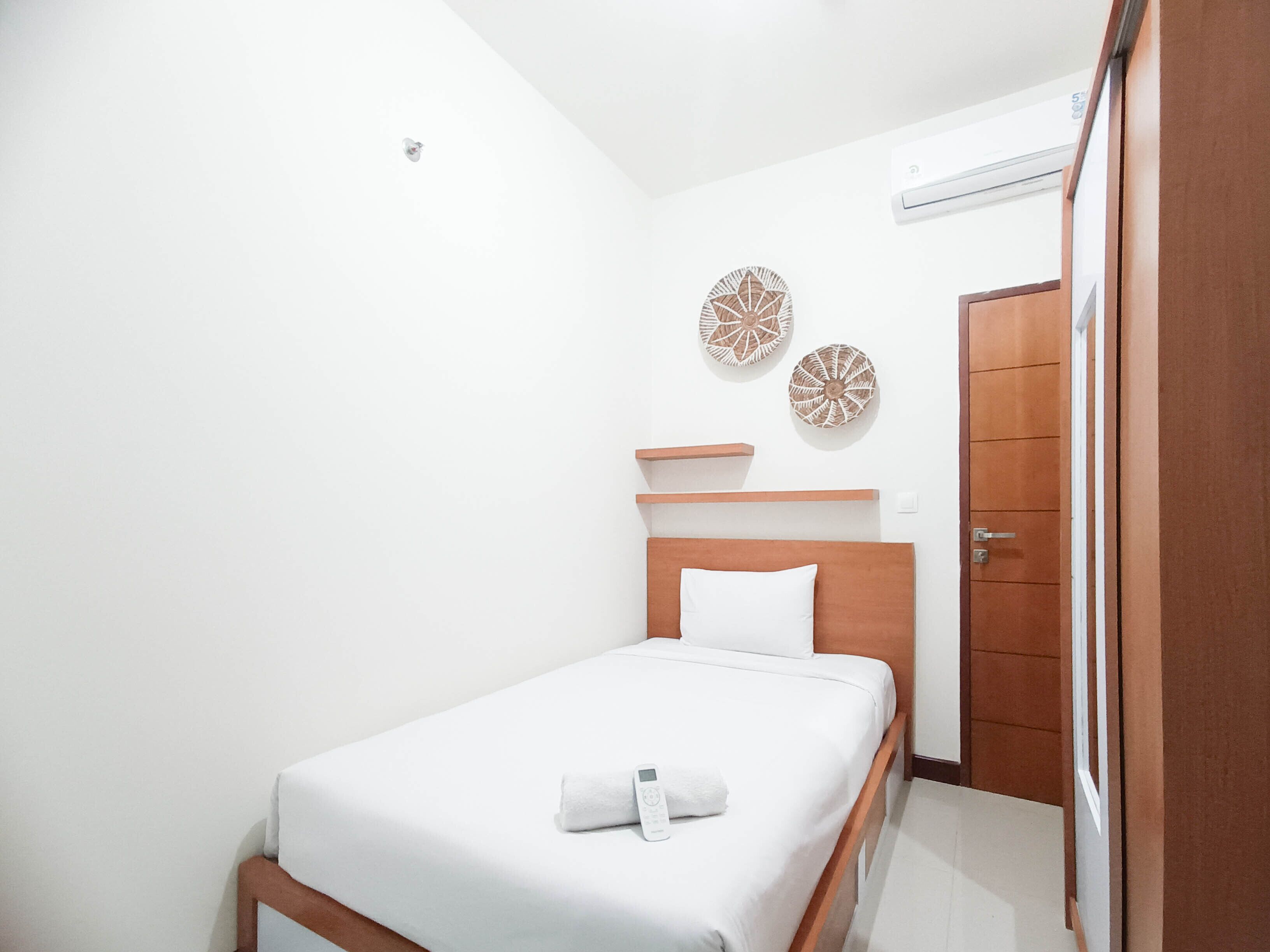 Una cama pequeña y ordenada, con colchón blanco y cabecera de madera. Dos estantes fijos en la pared se encuentran encima de la cama, y una puerta de madera a la derecha.