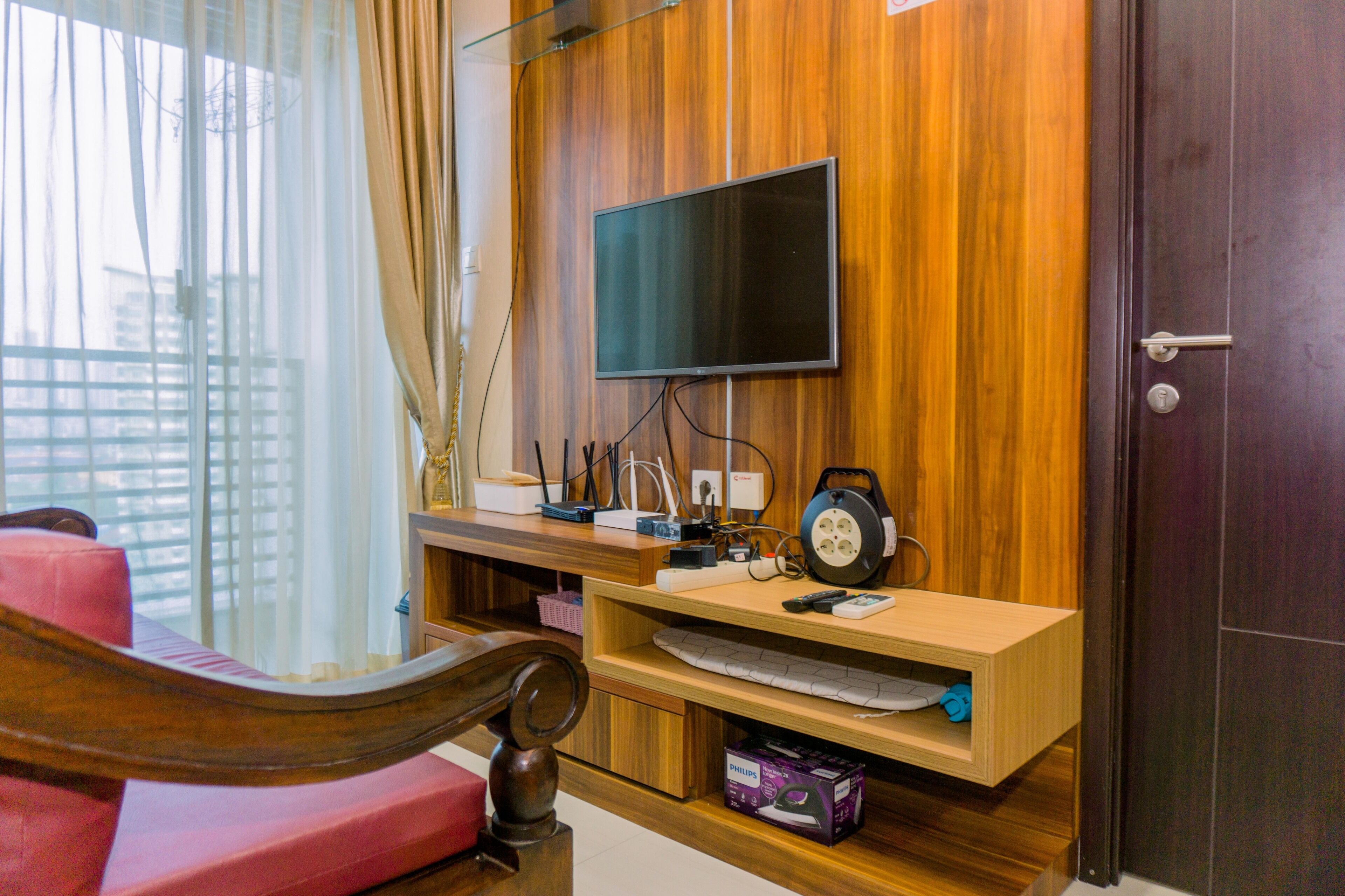 Een kamer met een houten tv aan de muur, een bureau met een computer en een rode stoel.