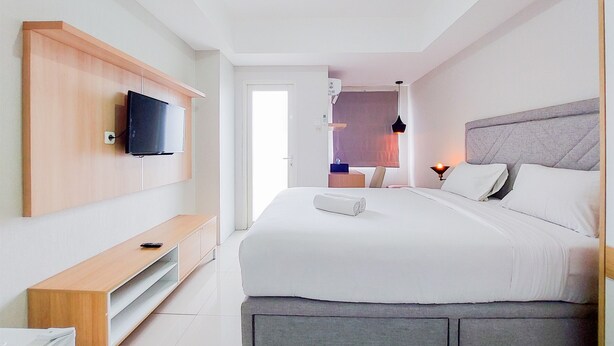 A modern hotel room with a large bed, a flat-screen TV mounted on the wall, a bedside table, and a small desk area. - Cozy Living Studio Room At Baileys Apartment (South Tangerang)