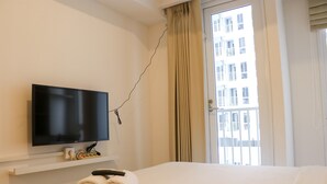 A hotel room with a bed, a flat-screen TV mounted on the wall, a pair of rolled towels on the bed, and a window with curtains. - Stylish Studio At Tokyo Riverside Pik 2 Apartment (Teluknaga)